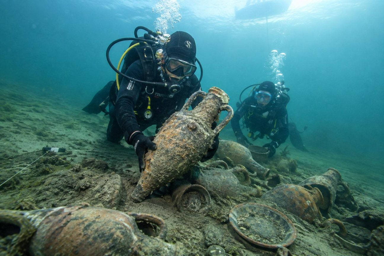 Plongeurs en équipement de plongée explorant le fond du lac de Neuchâtel parmi des amphores romaines et fragments de céramique enfouis dans la vase.