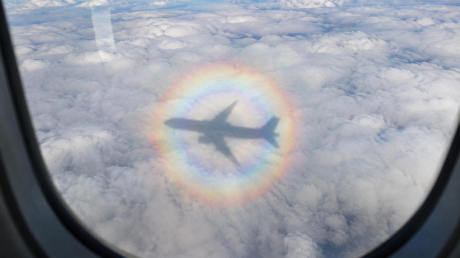 Halo multicolore irisé entourant l'ombre d'un avion projetée sur une couche de nuages blancs, visible depuis un hublot avec le soleil en arrière-plan