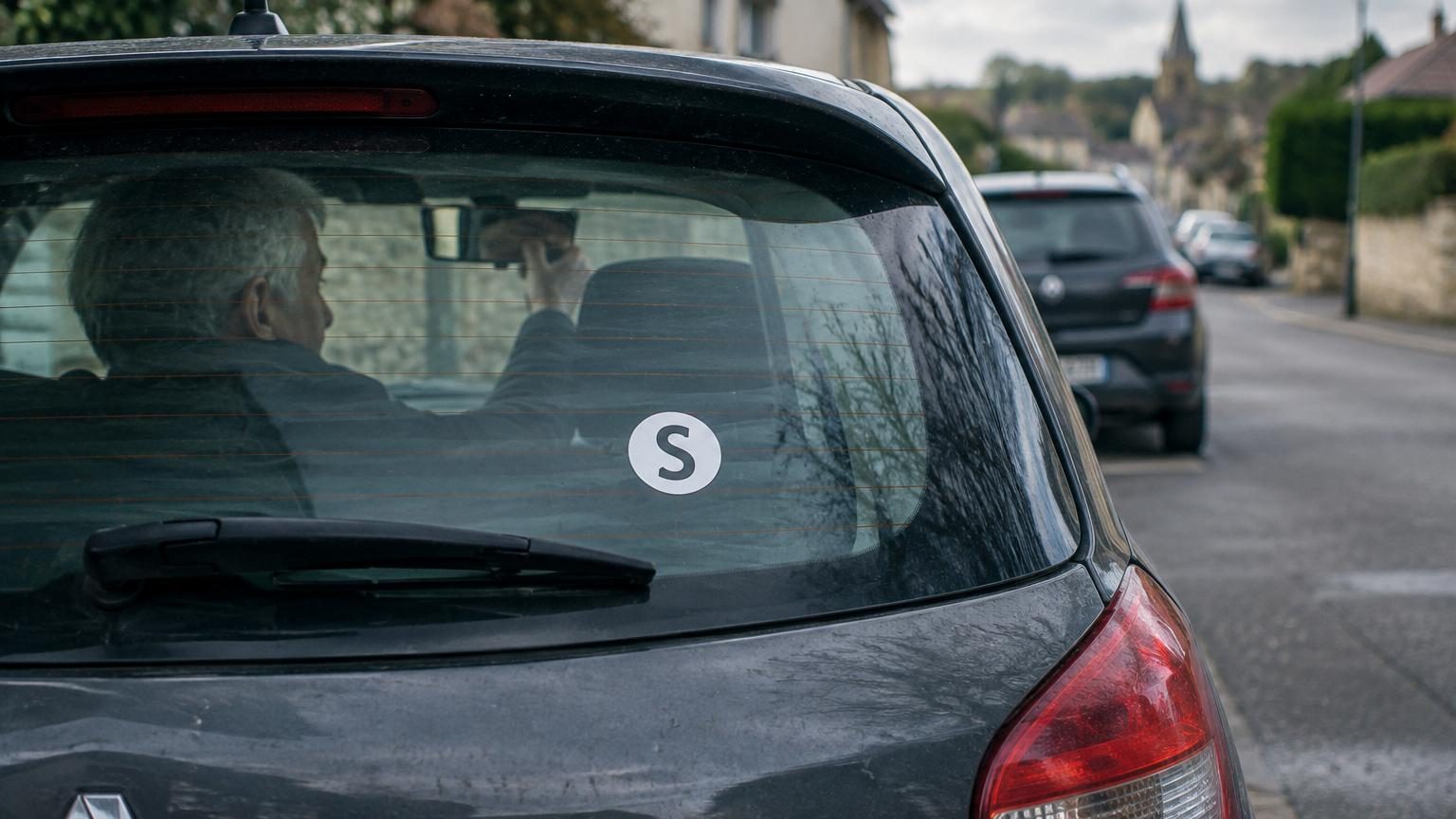 Pare-brise de voiture avec un autocollant représentant la lettre S en blanc sur fond rouge ou orange, symbole des conducteurs seniors.