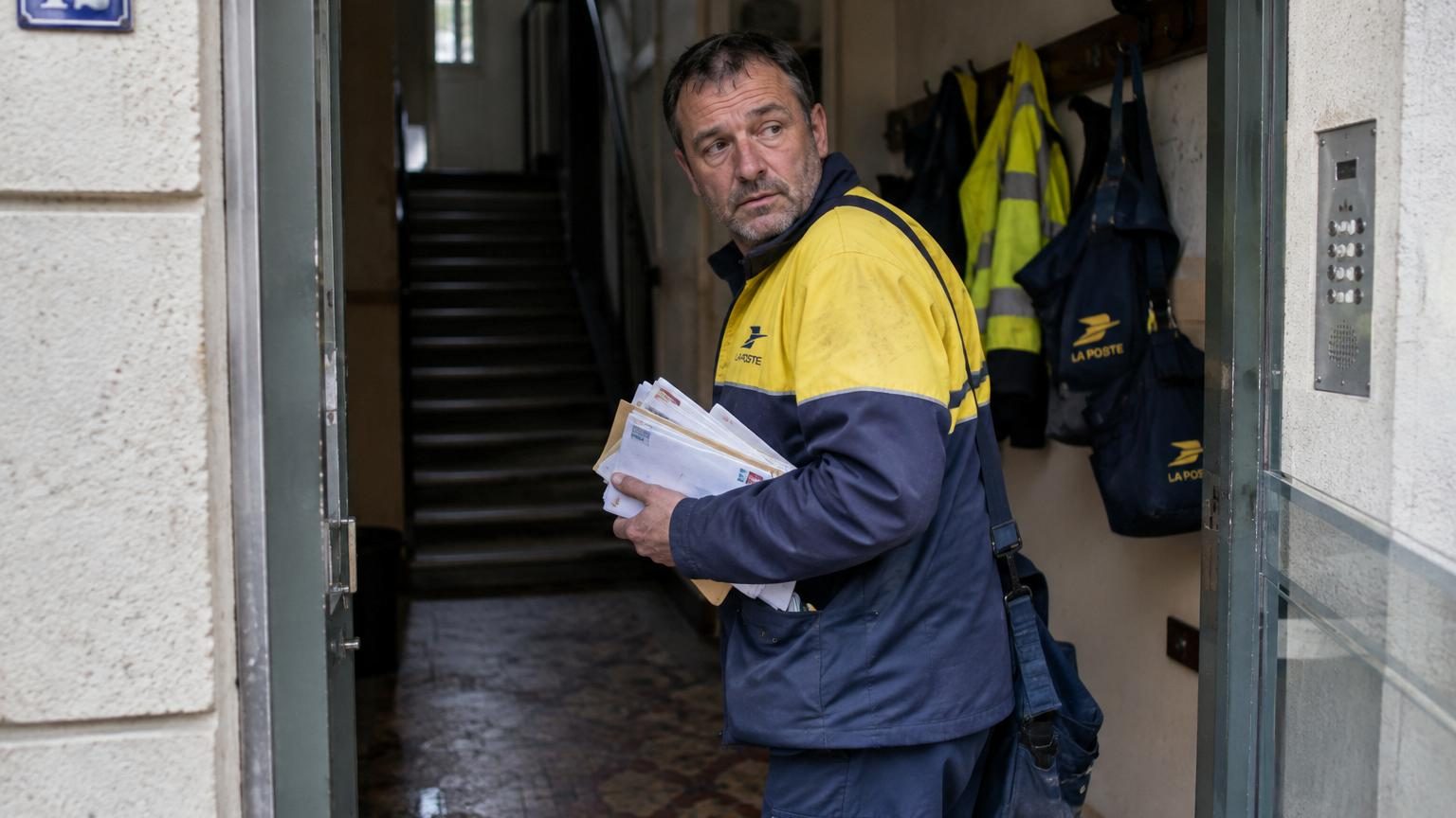 Un facteur chargé de colis sur son épaule, prêt à partir en tournée depuis le parking du centre de tri, illustrant la réalité quotidienne fragmentée de ce métier.