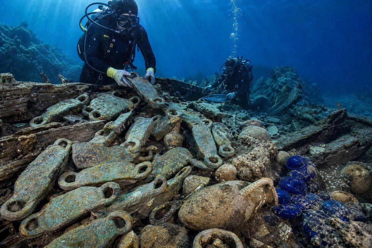 Vue sous-marine d'une épave antique avec des lingots de cuivre en peau de bœuf dispersés sur le fond rocheux entouré de sédiments et de débris.