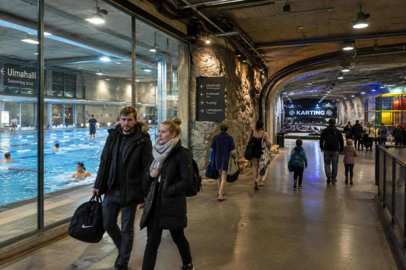 Vue intérieure d'une piscine spectaculaire creusée dans le granit, avec carrelage coloré lavande, jaune et bordeaux, formes géométriques suspendues au plafond et parois rocheuses visibles.